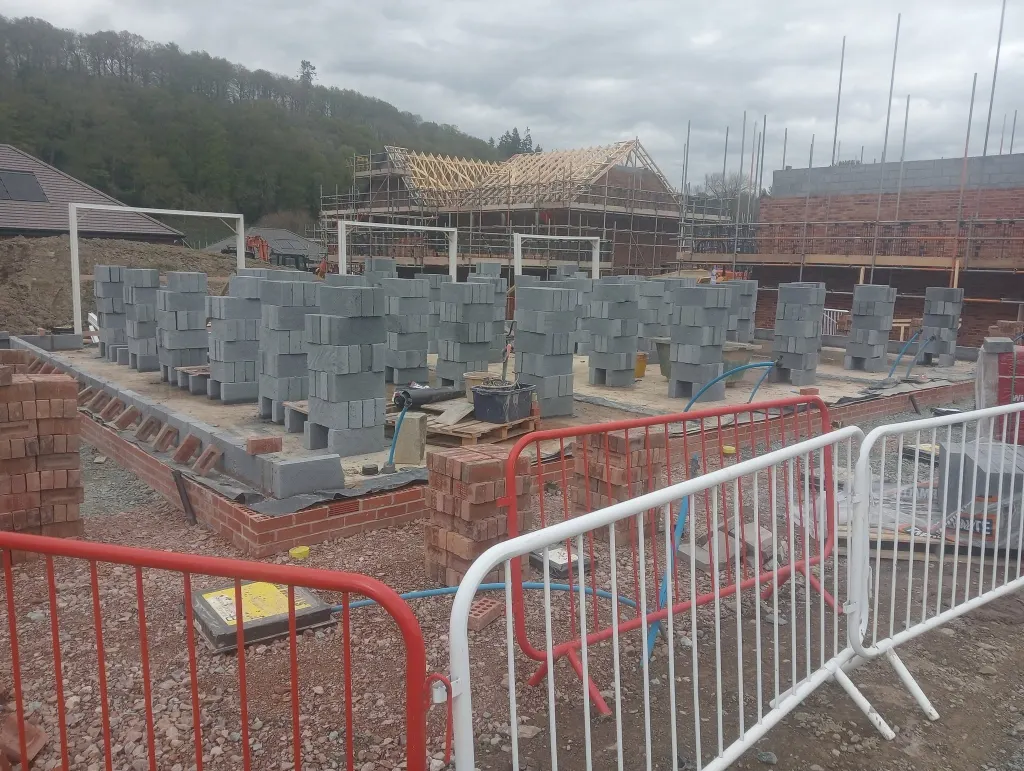 Beam-and-block ground floors across several plots with grey blocks stacked ready, scaffold and roof trusses behind