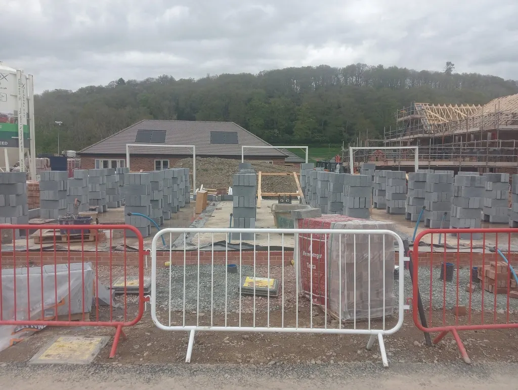 Grey concrete blocks stacked on a beam-and-block floor behind site barriers, with a finished bungalow beyond