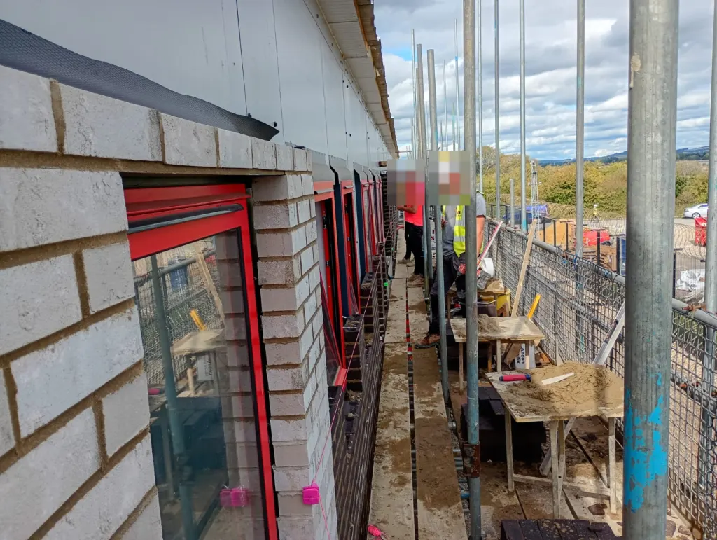Bricklayers working off scaffold on a white-grey brick wall with red-framed windows, spot boards and mortar ready