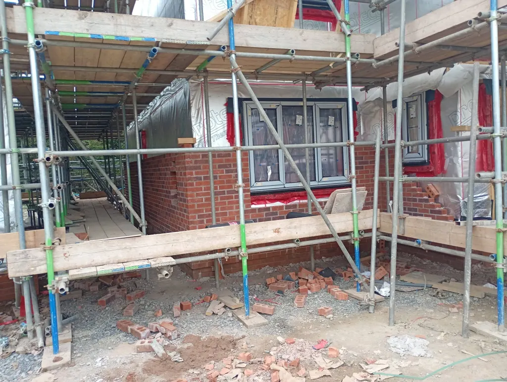 Facing brick under a wide window with red cavity closers, framed by scaffold and broken brick cuts on the ground