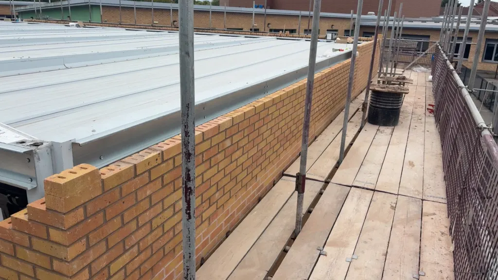 Top of a buff facing brick wall seen from the scaffold, beside a white profiled metal roof, with another building below