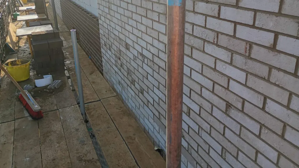 Run of buff facing brick wall with a darker engineering brick course near the base, seen from the scaffold