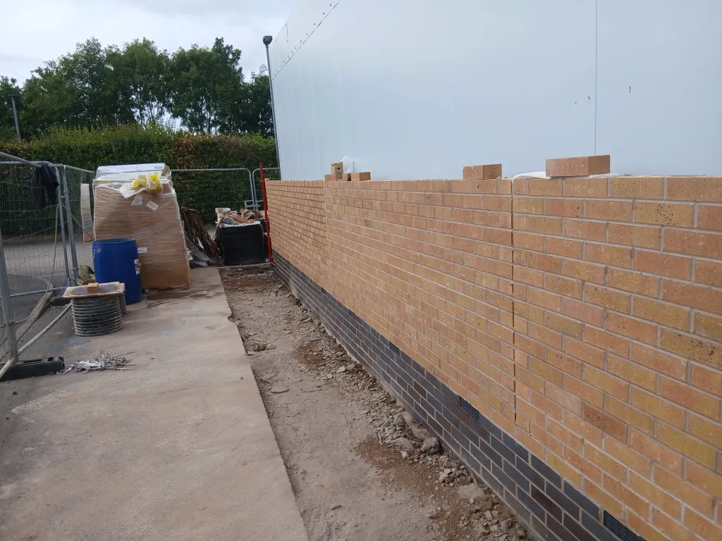 Buff facing brick wall racked back along a white-clad building, with materials, a drum and fencing at the end