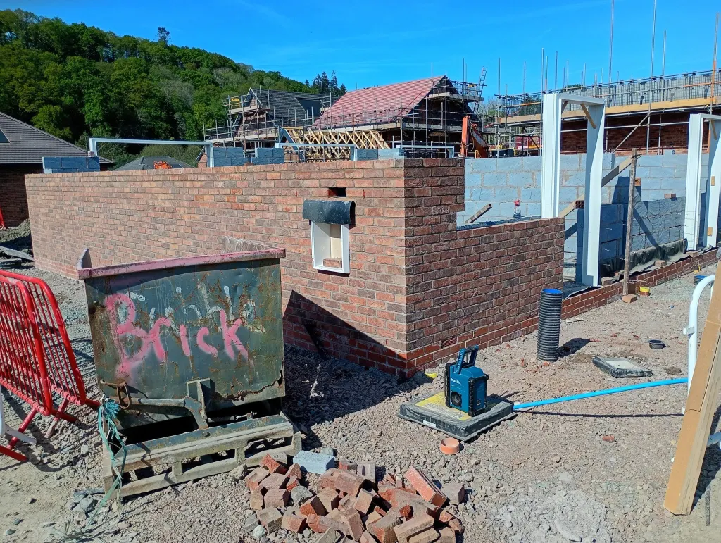 Bungalow walls in red facing brick with a meter box and a skip marked Brick, with scaffolded houses in the distance