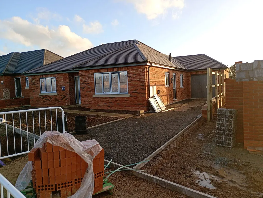 Completed bungalow in red facing brick with a hipped grey tiled roof, garage alongside and wrapped brick pack in front