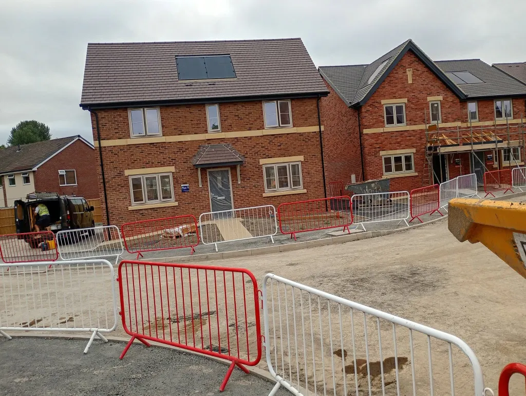 Detached two-storey house in red brick with a buff stone band and porch canopy, fenced off from the new estate road