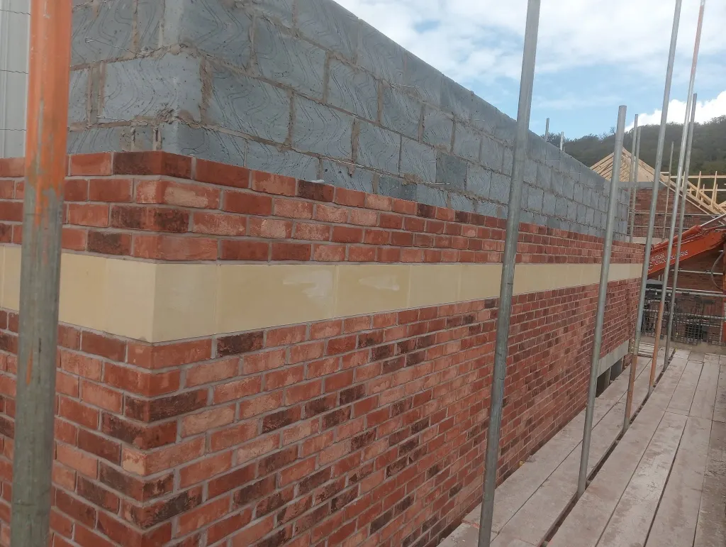 Buff stone band course running through red facing brick, with grey blockwork above and scaffold standards alongside