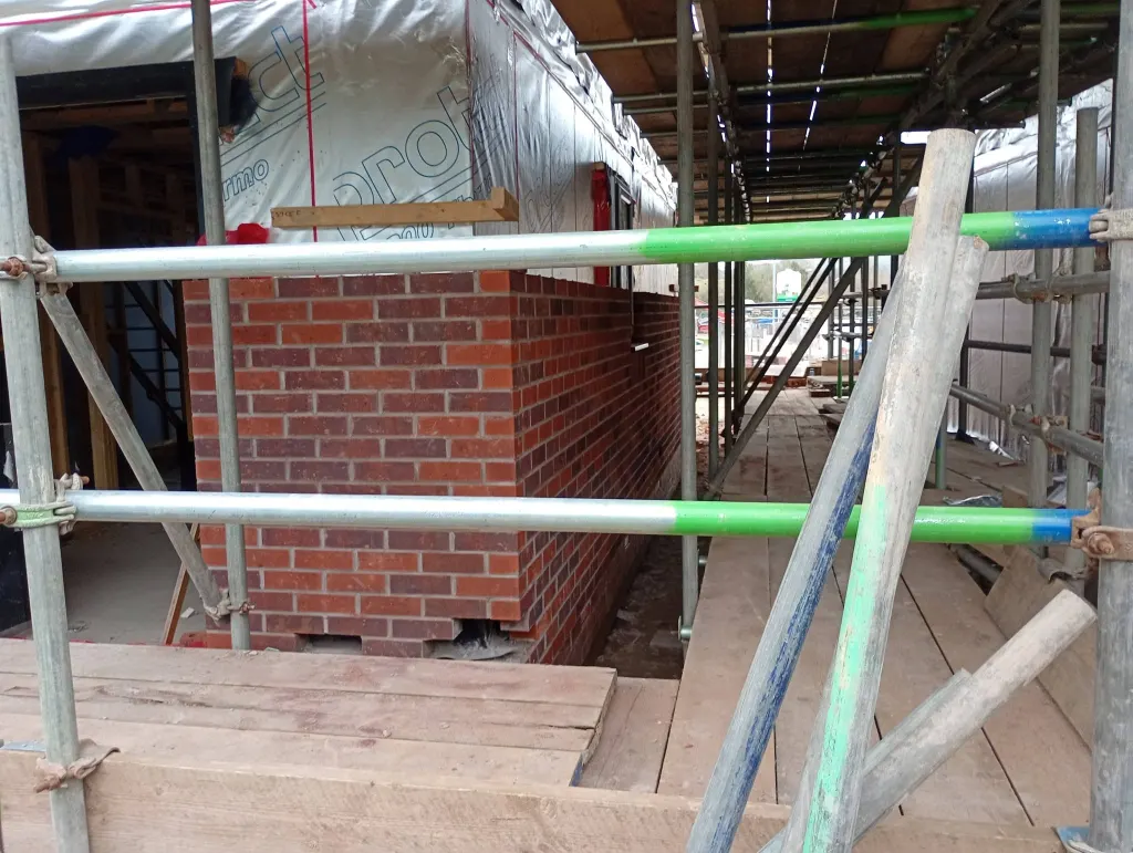 Short return of red facing brick beside a scaffold walkway, with membrane-covered framing above and a door opening
