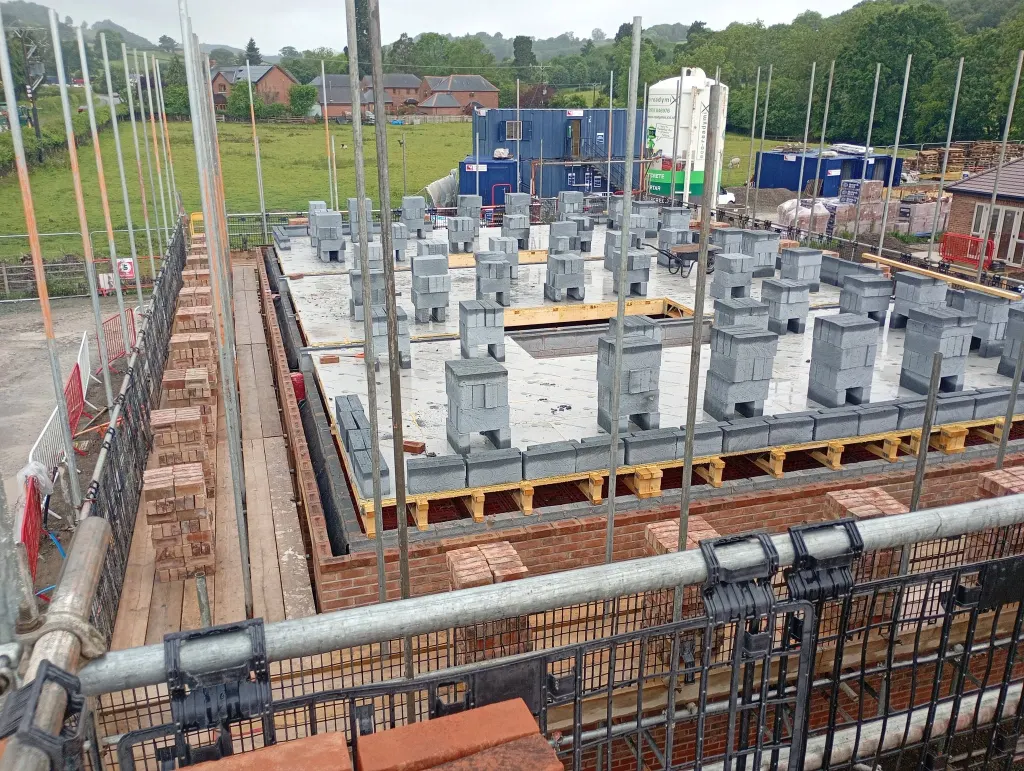 Blocks stacked across a floor deck inside a facing brick outer leaf, seen from scaffold with site cabins behind