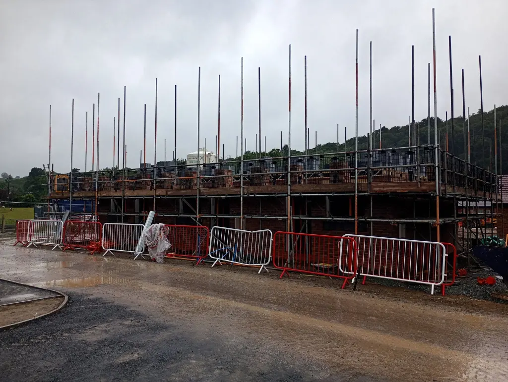 Row of plots wrapped in scaffold with brickwork up to first floor, barriers along a wet site road and trees behind