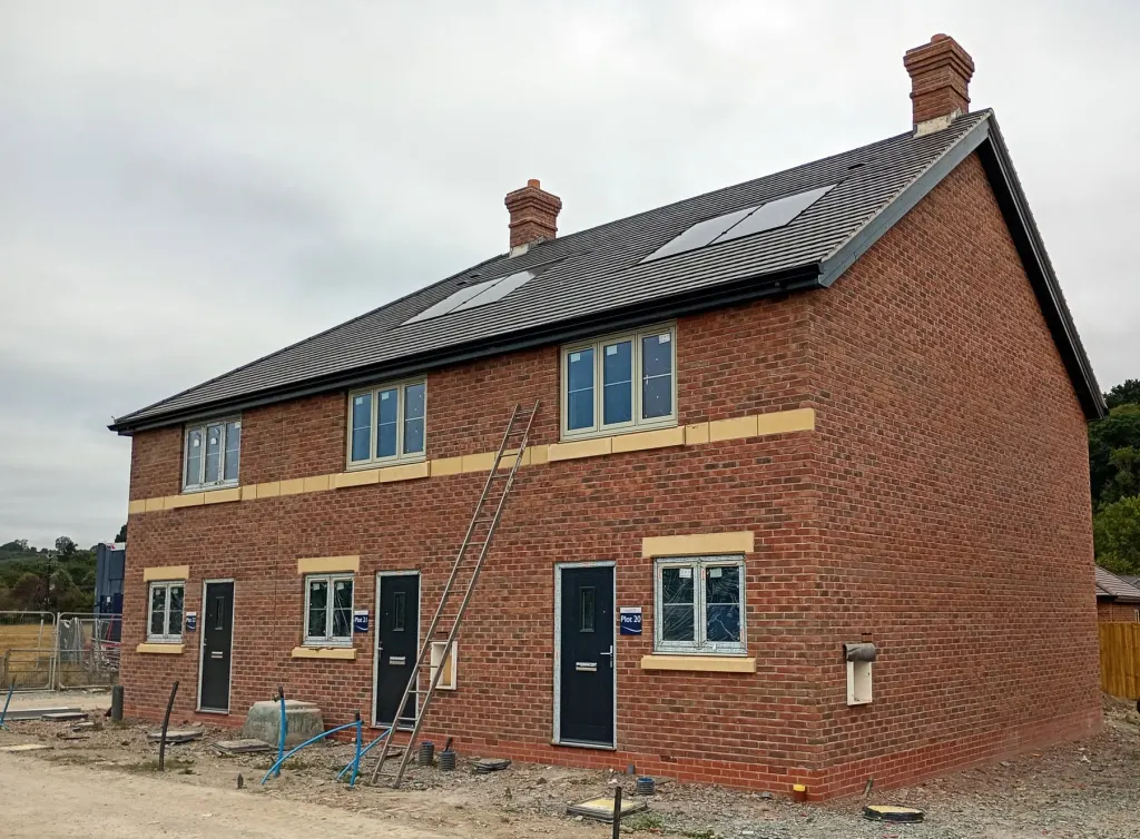 Semi-detached pair in red facing brick with two chimneys, roof windows, a buff stone band and grey front doors