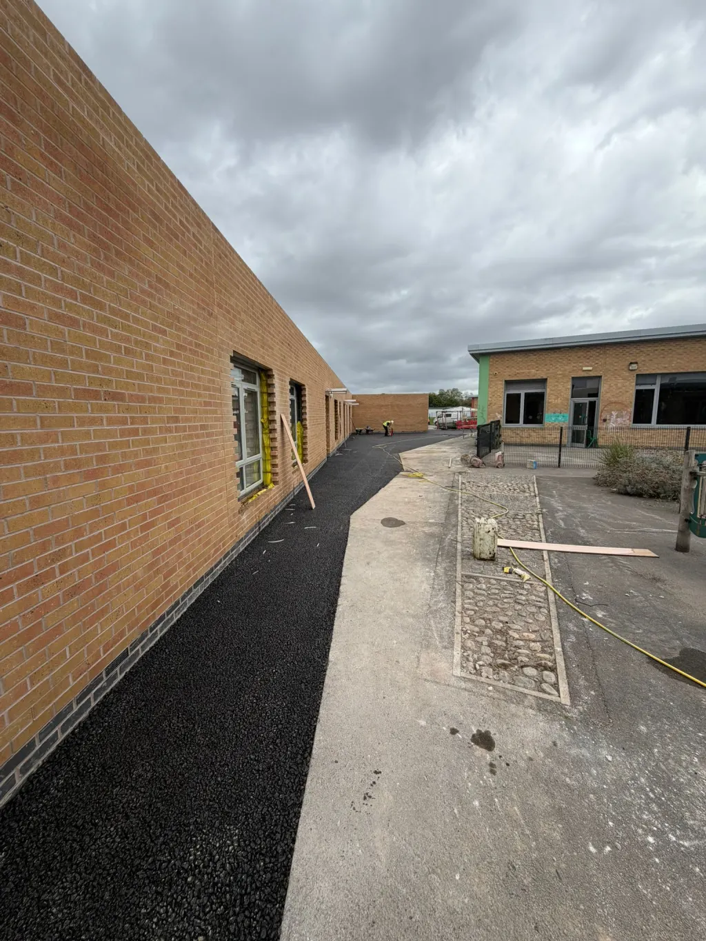 Long single-storey building in buff facing brick with a dark engineering brick base, beside a new tarmac footpath