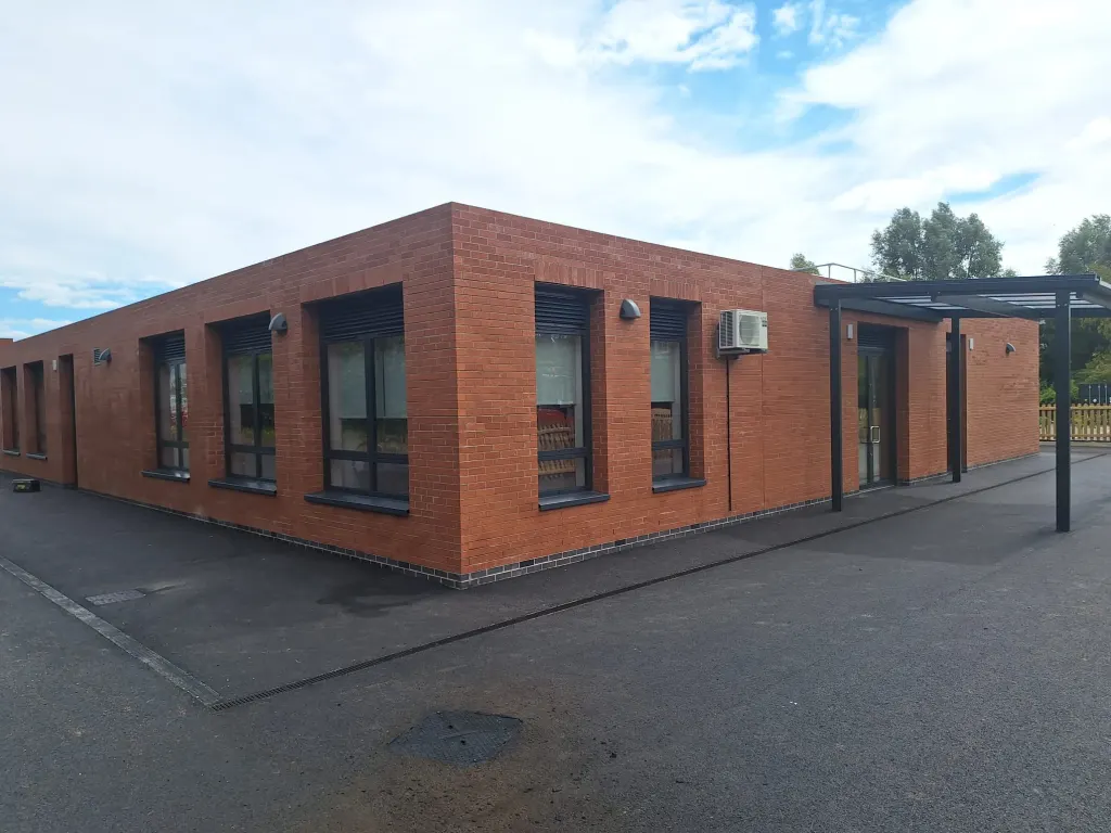 Single-storey building in red facing brick with dark window frames and a steel entrance canopy beside a tarmac yard