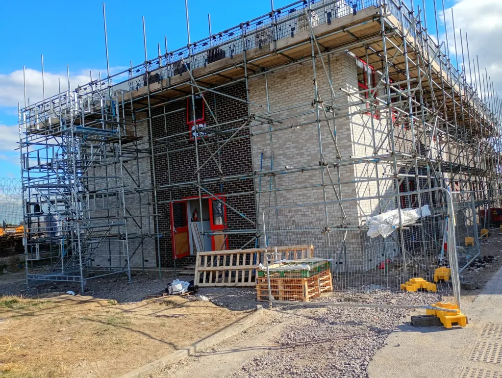 Two-storey building in white-grey and dark facing brick under scaffold with a stair tower and red-framed doors
