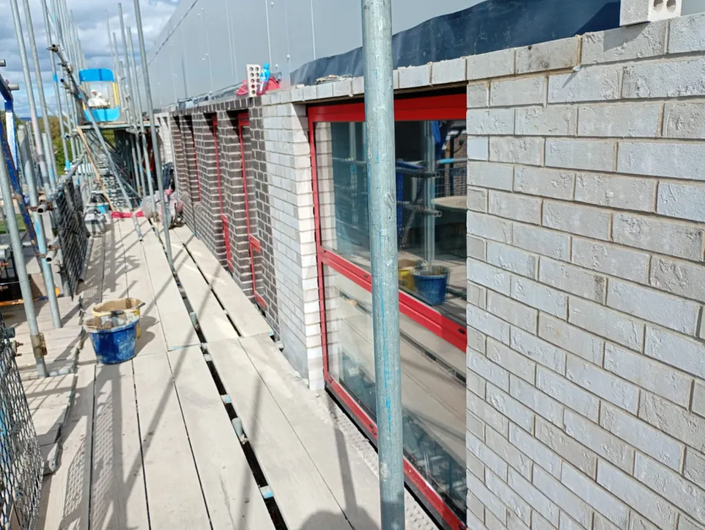 White-grey facing brick around red-framed windows, with dark brick panels and scaffold boards along the elevation