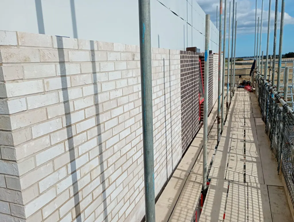 White-grey facing brick with a dark brick panel, seen along a scaffold walkway with open countryside beyond